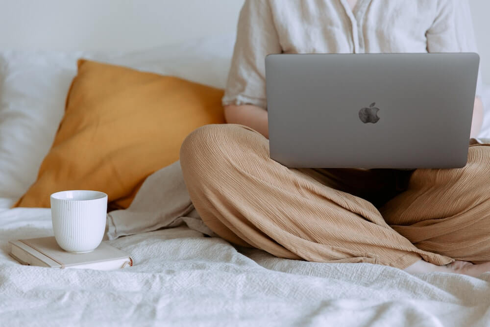 Person wearing flowy tan pants and white shirt sits crosslegged on bed with linen sheets, with a laptop in their lap and a coffee mug next to them