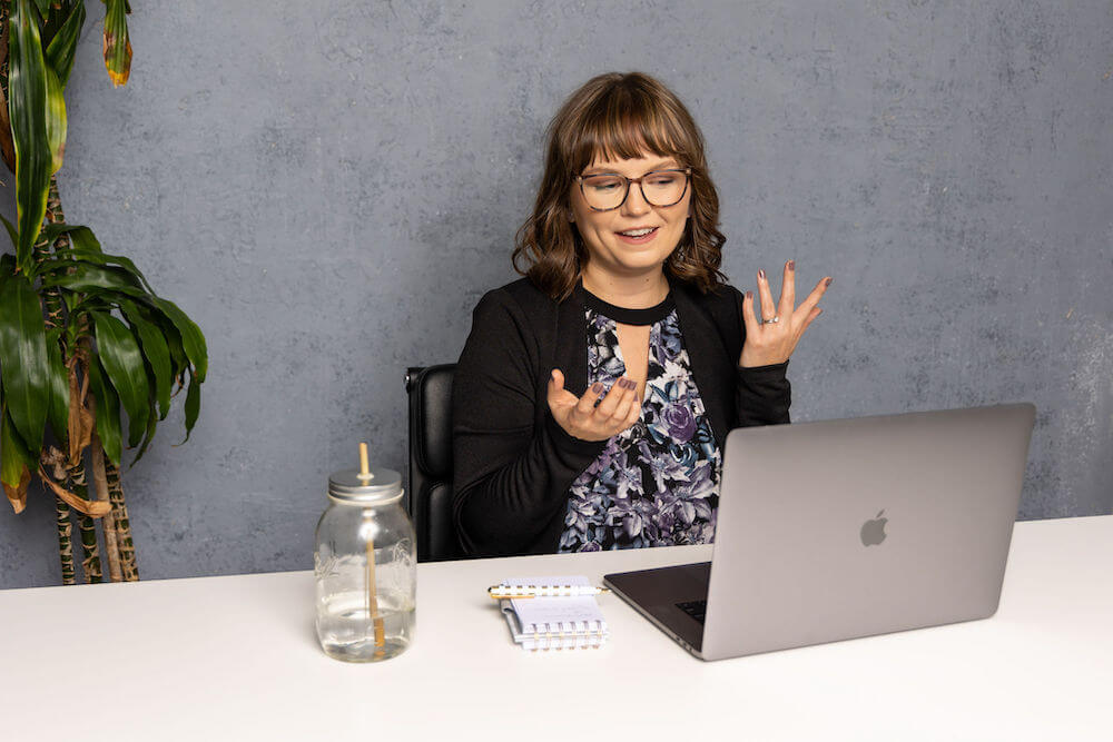 Lucy sitting in front of a gray backdrop at a white table. She appears to be on a video coaching session with a career coaching client, as she's mid-sentence and gesturing with her hands in the air while looking at her MacBook screen. She wears a purple floral top and black blazer.