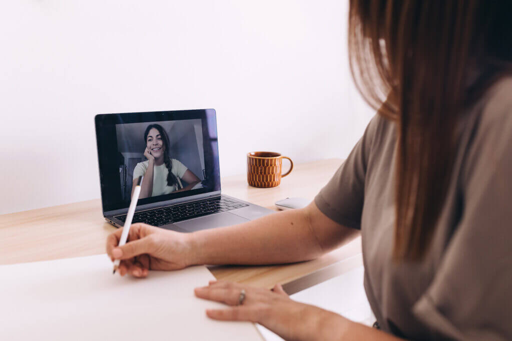 Feminine-presenting person with long brunette hair and loose gray shirt sits at a light-colored wood desk, holding white pencil to blank paper; they're on a video conference with another feminine-presenting person shown on a laptop screen