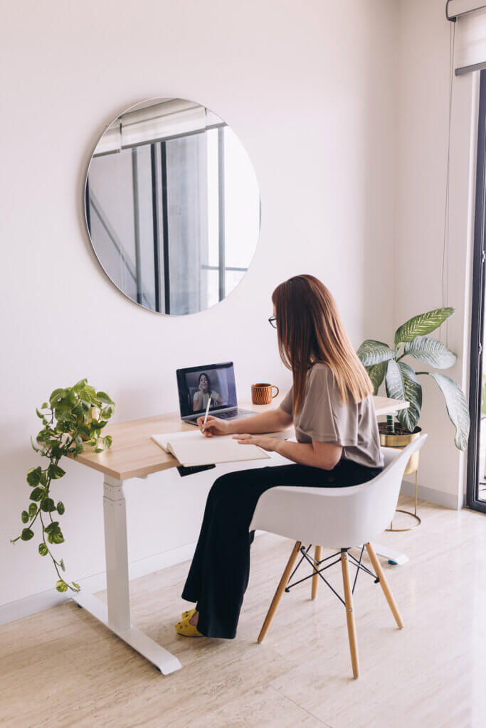 A feminine-presenting person with long light brown hair sits in a bright white workspace in a white chair with white pine desk, plants, and a round mirror. She appears to be on a video chat, maybe with a career coach, and is taking notes in a notebook.