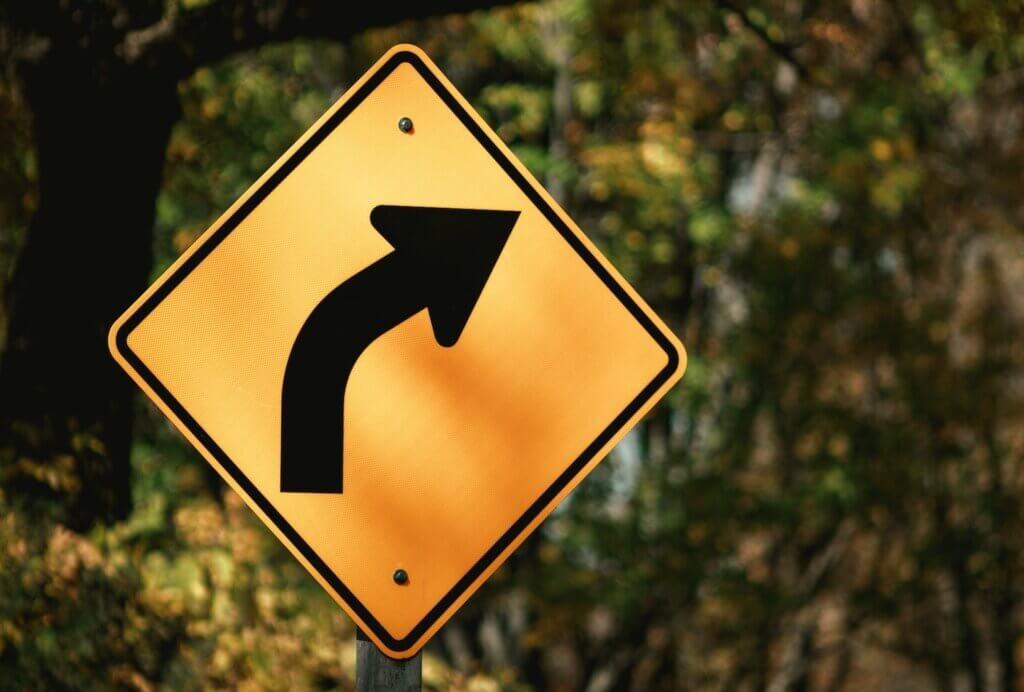 A partially shaded traffic sign shows a curved arrow to indicate a bend in the road, which is used as a metaphor for changing careers. In the background, trees with green and yellow foliage are out of focus