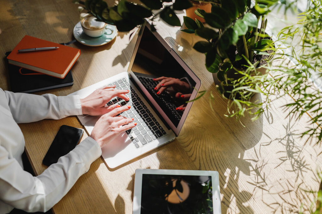 Feminine-presenting hands with orange fingernails type on a MacBook. The laptop sits on a woodgrain counter in the sunshine, surrounding by plants, a stack of notebooks, and a tablet.