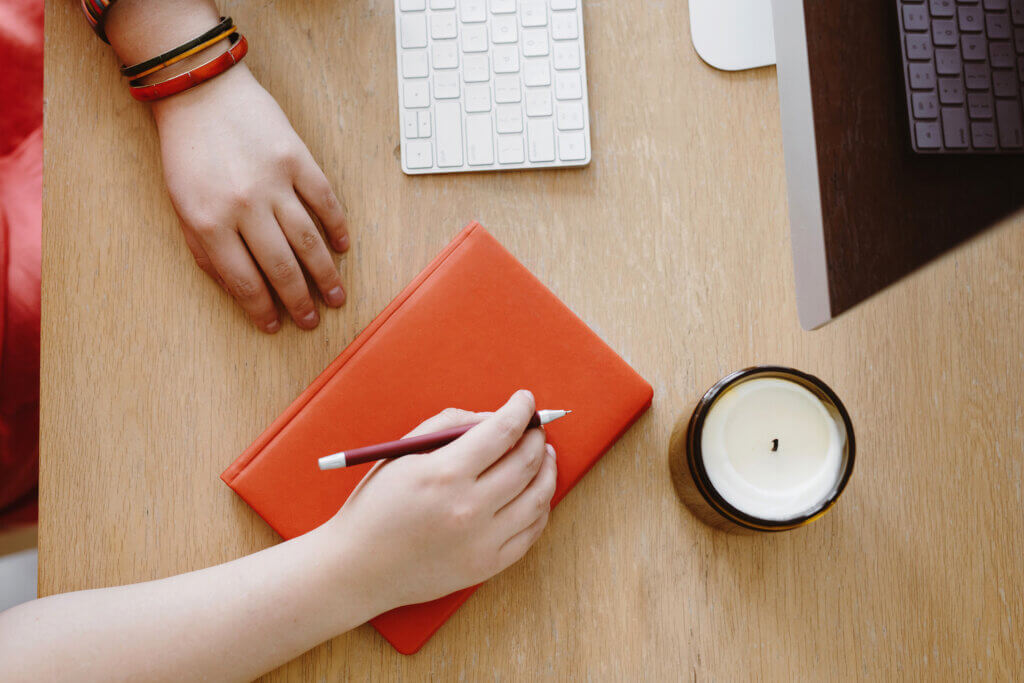 An overhead view of a light woodgrain desktop. A bright coral orange notebook sits in the center. Hands that appear to belong to a light-skinned white woman rest on the desktop: one on the desk itself and the other holding a pen and resting on top of the orange notebook. A partly burned candle sits near the notebook along with the edge of a white Apple keyboard and desktop monitor