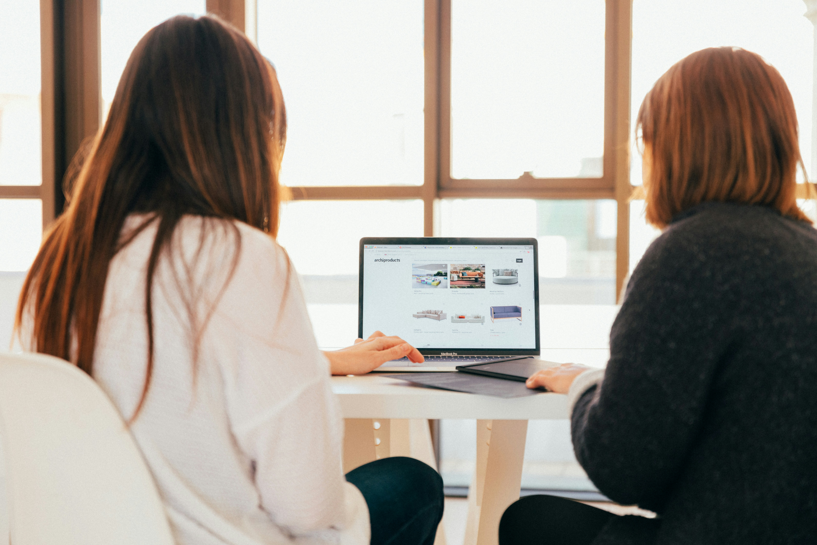 Two feminine-presenting, likely white people sit facing away from the camera. They're both looking at a laptop on a white table as though collaborating on something, like career discovery counseling or coaching. 