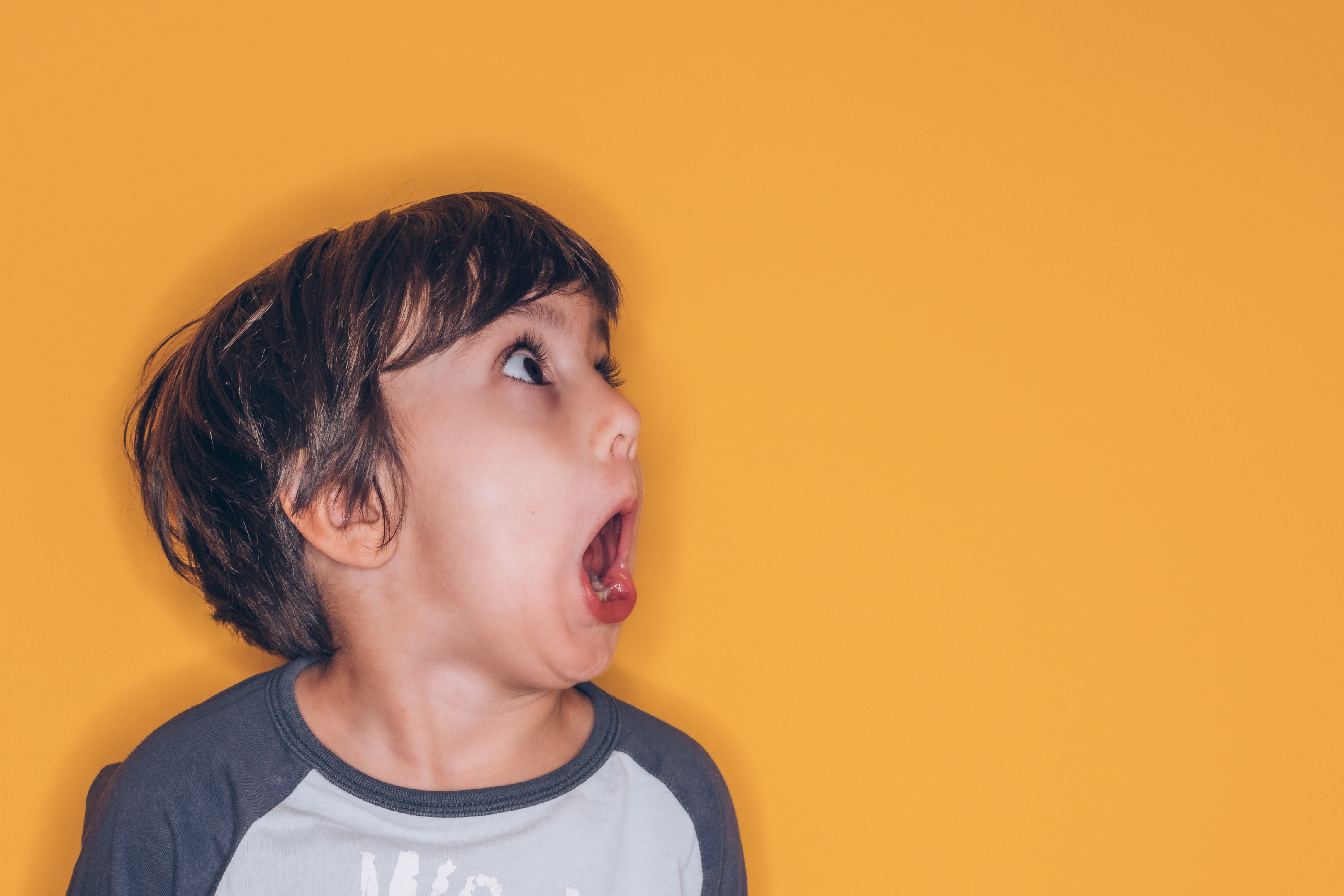 A child with medium skin tone and brown hair wears a blue baseball t-shirt and stands in front of a solild yellow backdrop. Their eyes and mouth are open wide and they look off-camera at something as though completely surprised or shocked — maybe by a career discovery epiphany.