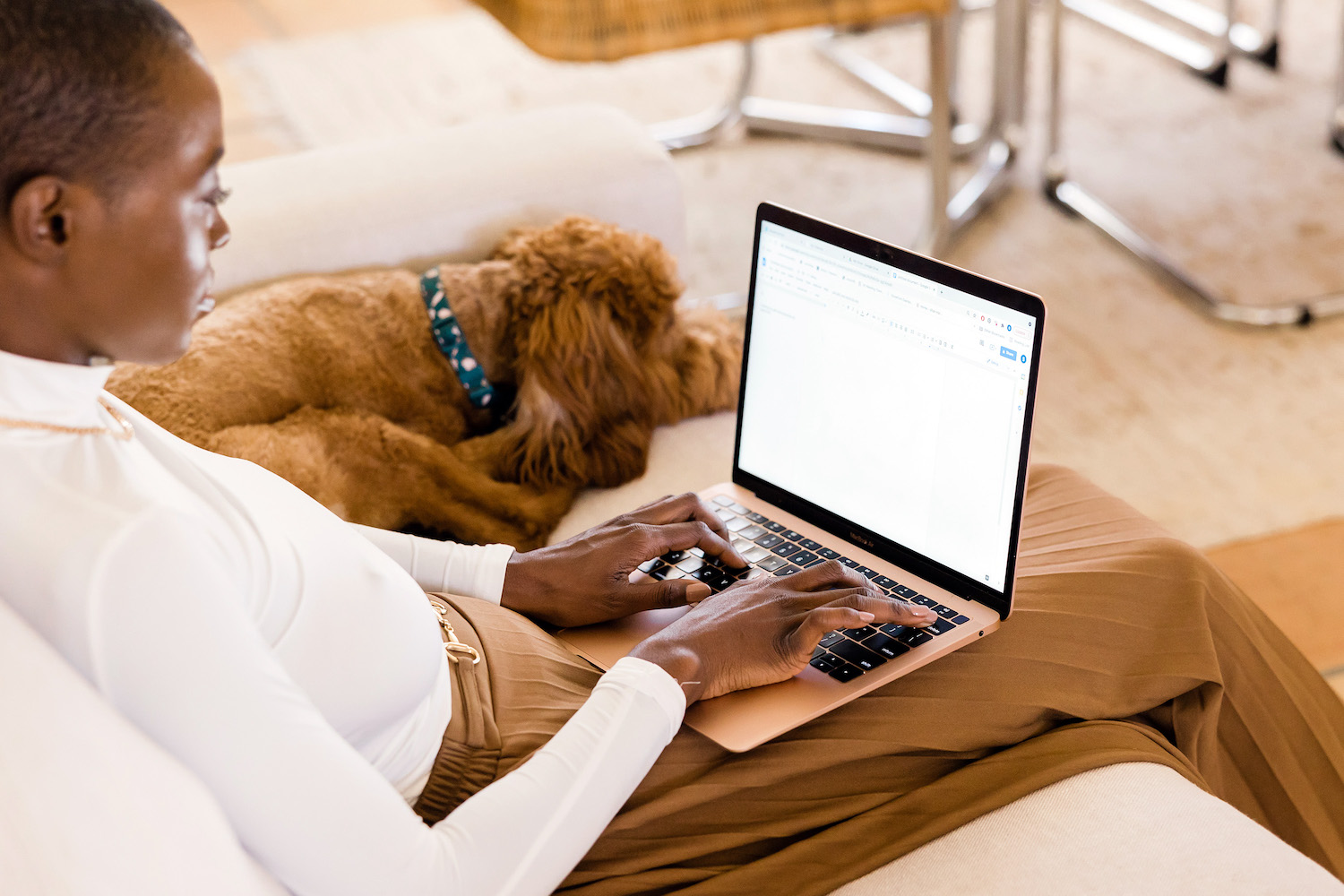 A Black feminine-presenting person sits on a couch with a rose gold Macbook on her lap. She's typing although we can't see what's on the screen. A golden poodle or doodle dog is next to her on the couch.