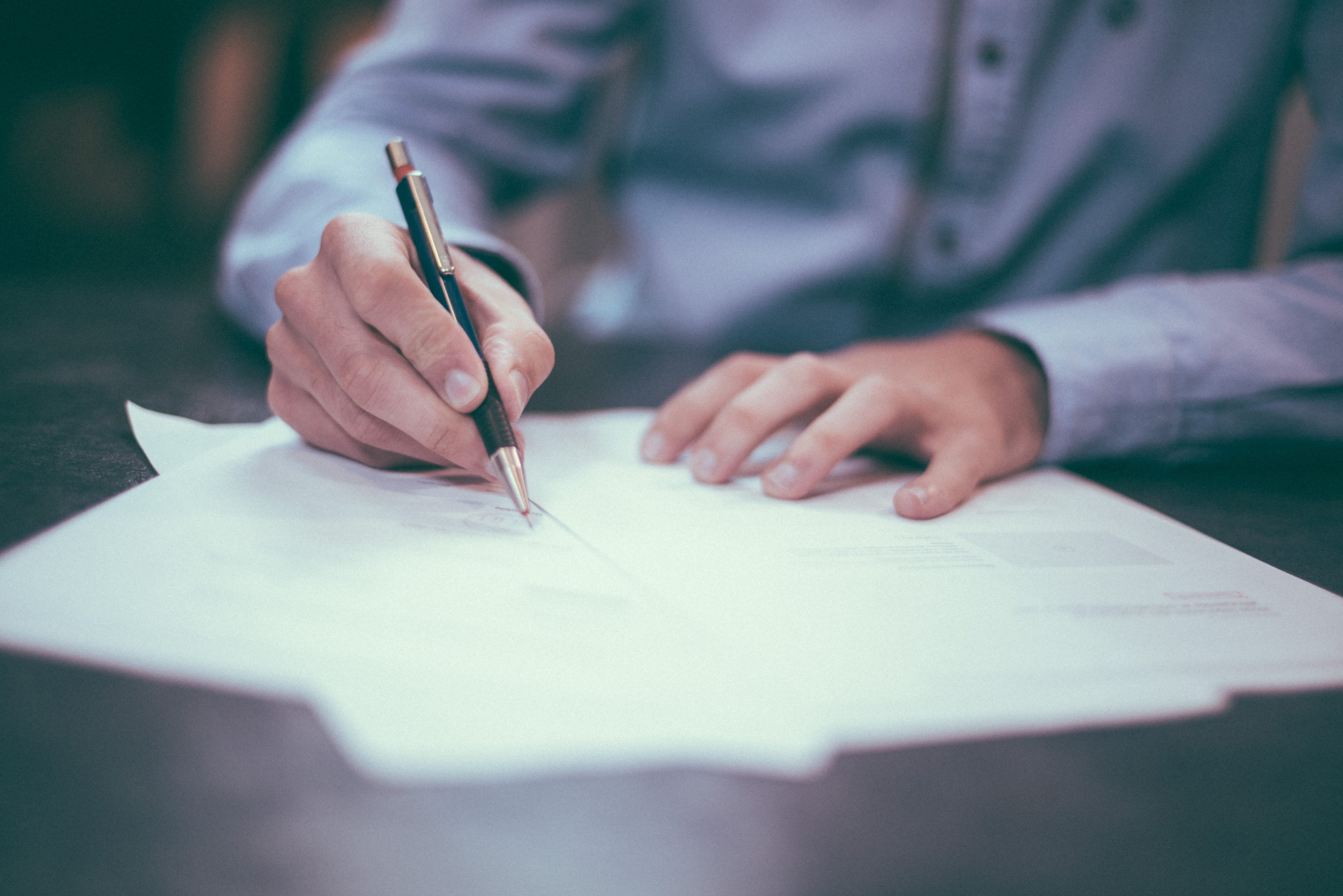 A focused image of masculine hands holding a pen and reviewing paper documents, like a career counselor reviewing a resume or cover letter. The person wears a light blue button down shirt with black buttons