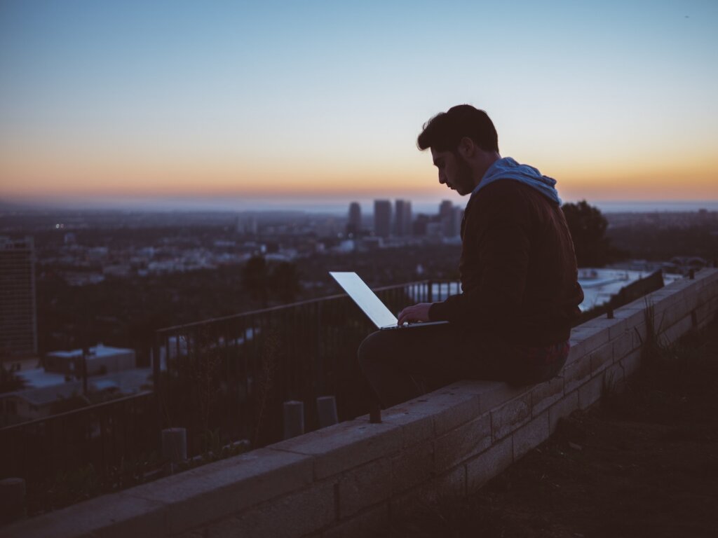 Masculine-presenting person sits perched on a ledge on top of a building, working on a laptop in his lap. It's dawn or dusk and a cityscape is out of focus in the background.