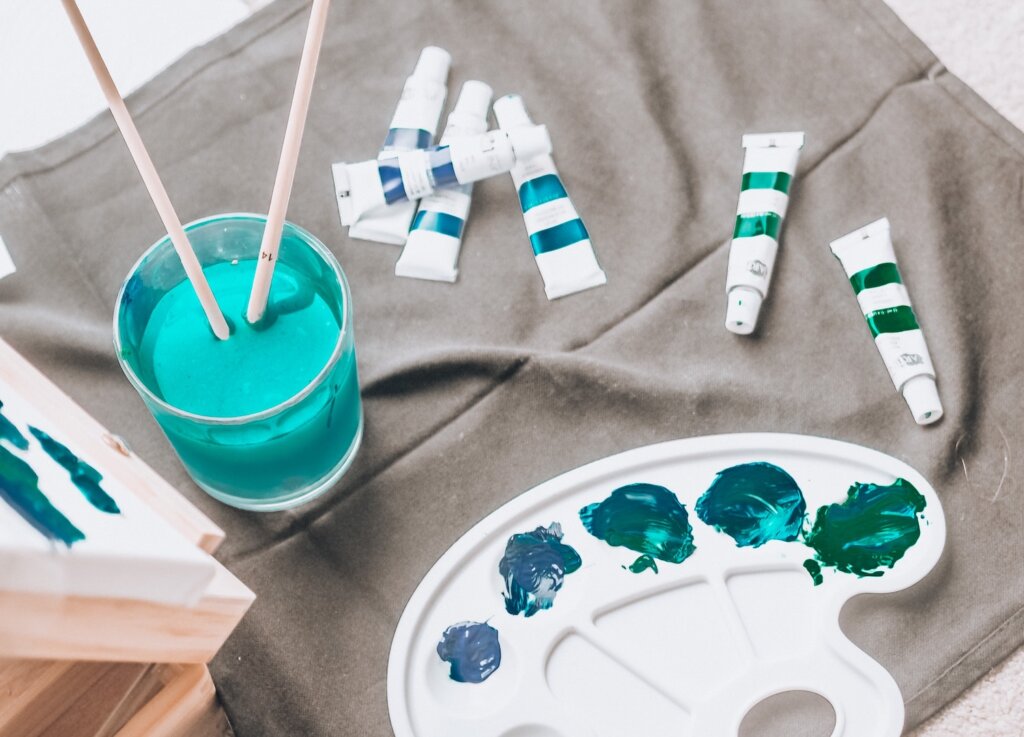White plastic paint palette on gray fabric. Paint brushes sit in a clear cup of bright teal paint water near an assortment of blue and green paint tubes, and the edge of a canvas is visible with some blue-green paint on it. This scene represents the fact that your existing skills and interests are applicable on your new career path.