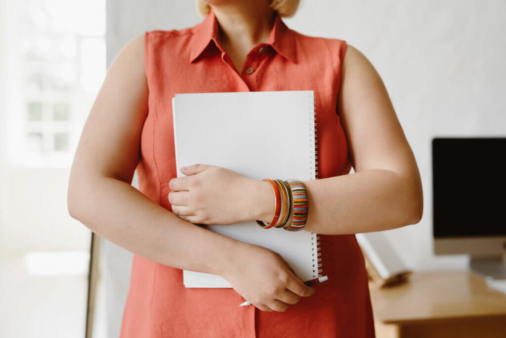 A white feminine-presenting person in a coral button-down sleeveless dress holds a blank spiral notebook to her chest. She has colorful bracelets on one wrist and holds a pen in the other hand.