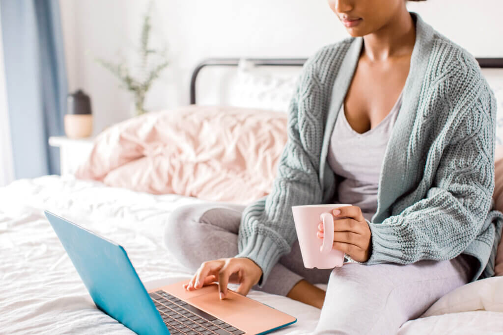 Black feminine-presenting person sits cross-legged on a bed with a pink mug in one hand and the other hand on a laptop near the trackpad. The bedroom is bright with white and pink accents.