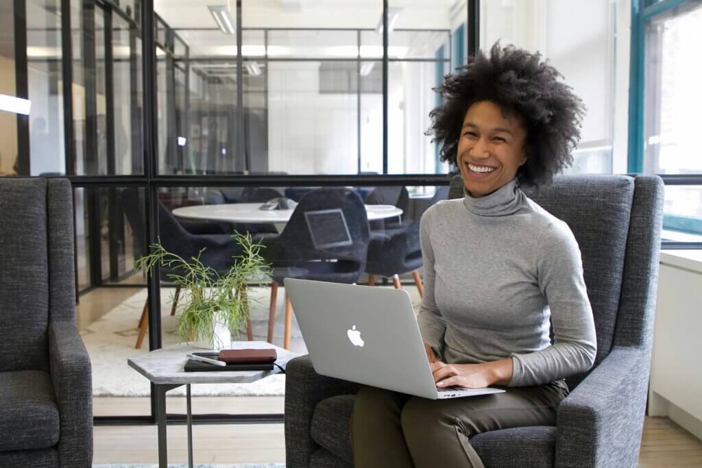 A Black feminine-presenting person with natural hair grins while sitting in an office with a laptop in their lap. They wear a light gray turtleneck and charcoal gray pants.