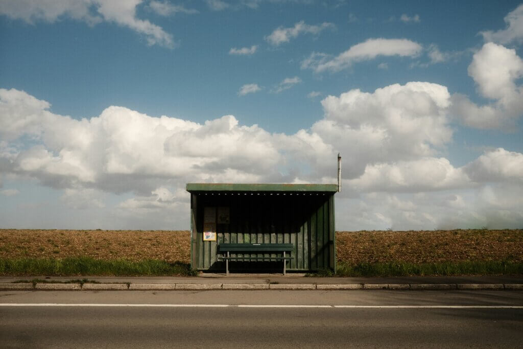 Blue sky with fluffy white clouds above a field. There's a road and an empty green shanty bus stop, which looks as though the bus doesn't come very often or at all. Kind of like waiting for your purpose or passion to come along, instead of playing an active role...