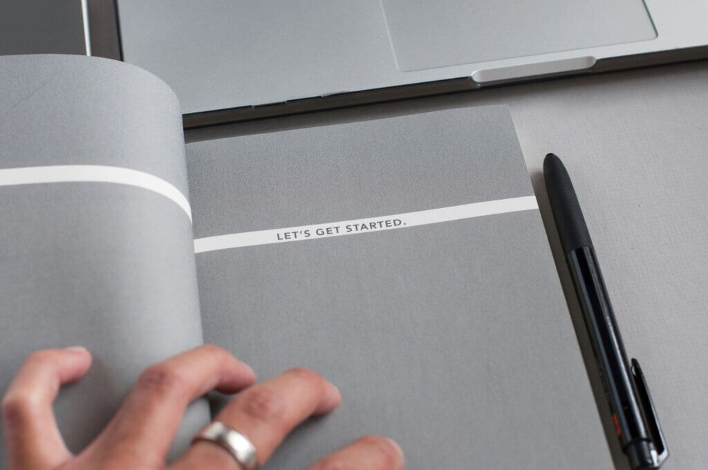 A hand with thick silver ring rests on an open notebook with dark gray pages. A white band spreading across the pages reads "LET'S GET STARTED." A black pen rests near the notebook and we also see the edge of a silver Macbook laptop.