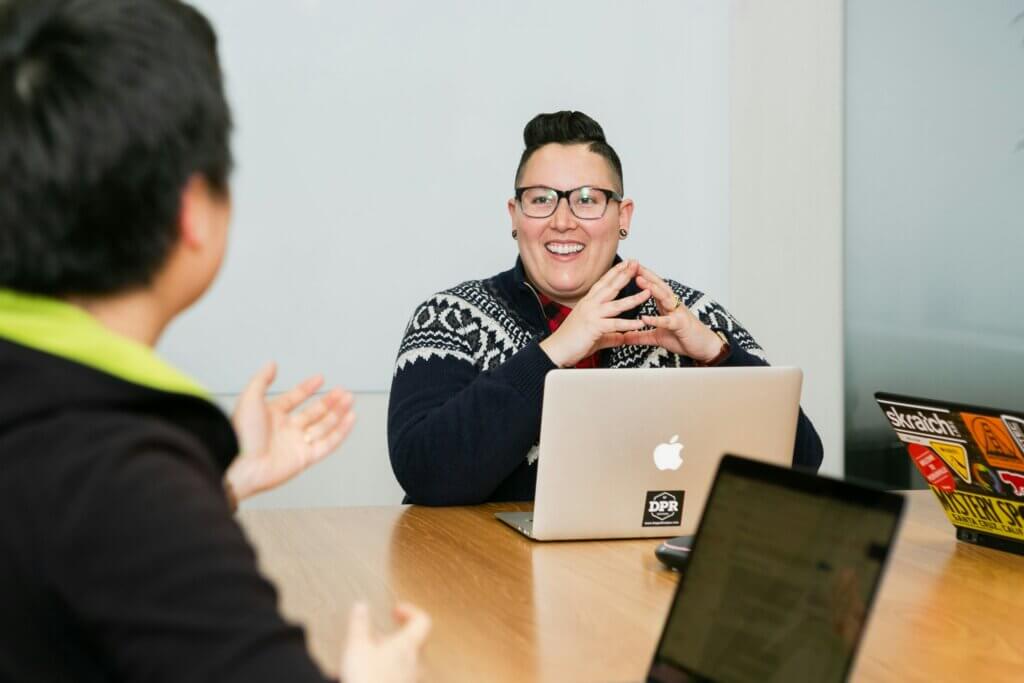Queer person with short brown hair, glasses, and a black sweater with white pattern sits at conference table with laptop in front of them, hands tented as they smile and talk to someone else at the table. 