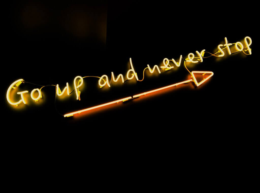 Black background with yellow-orange neon sign that says "Go up and never stop" with an arrow underlining the text