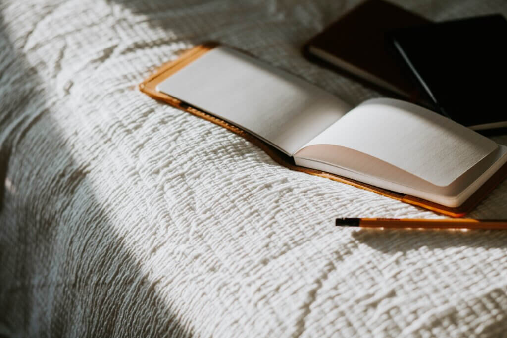 An open notebook on a white linen bedspread with a yellow pencil nearby; a strong shadow eclipses the scene so that the sun highlights the notebook and pencil.