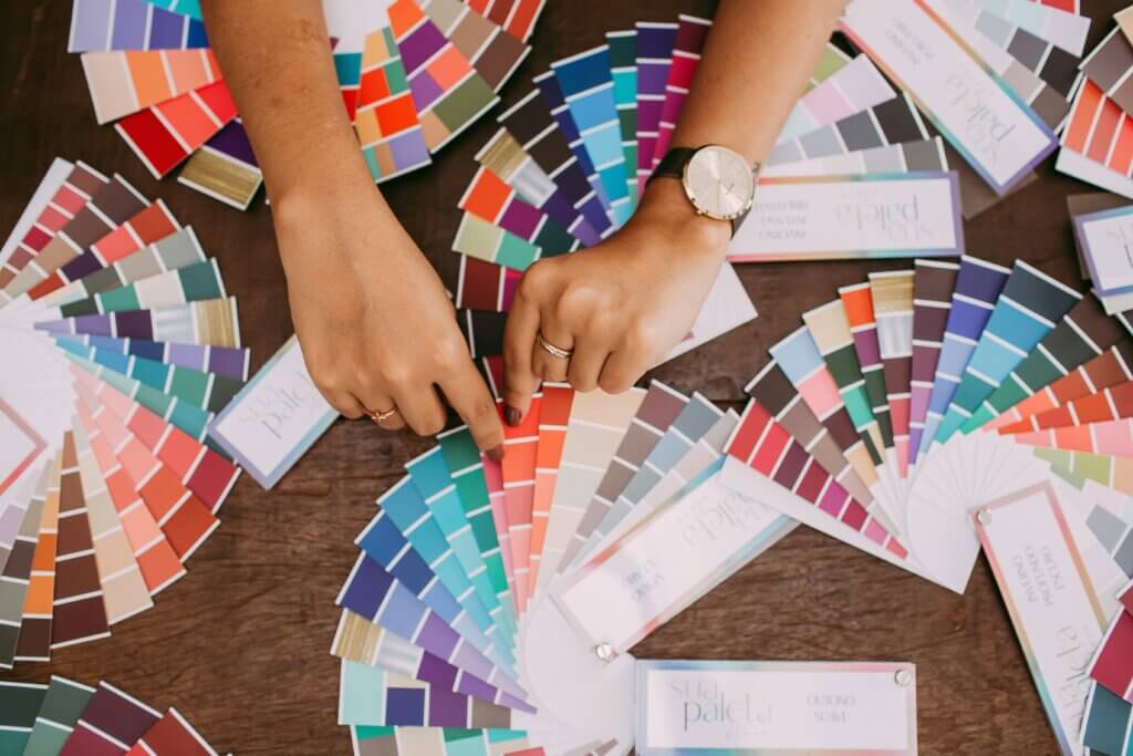 The feminine-presenting hands of a person of color with brown nail polish and rings hold a long, thin booklet of color palettes. On the table are several other versions of the same color palette.