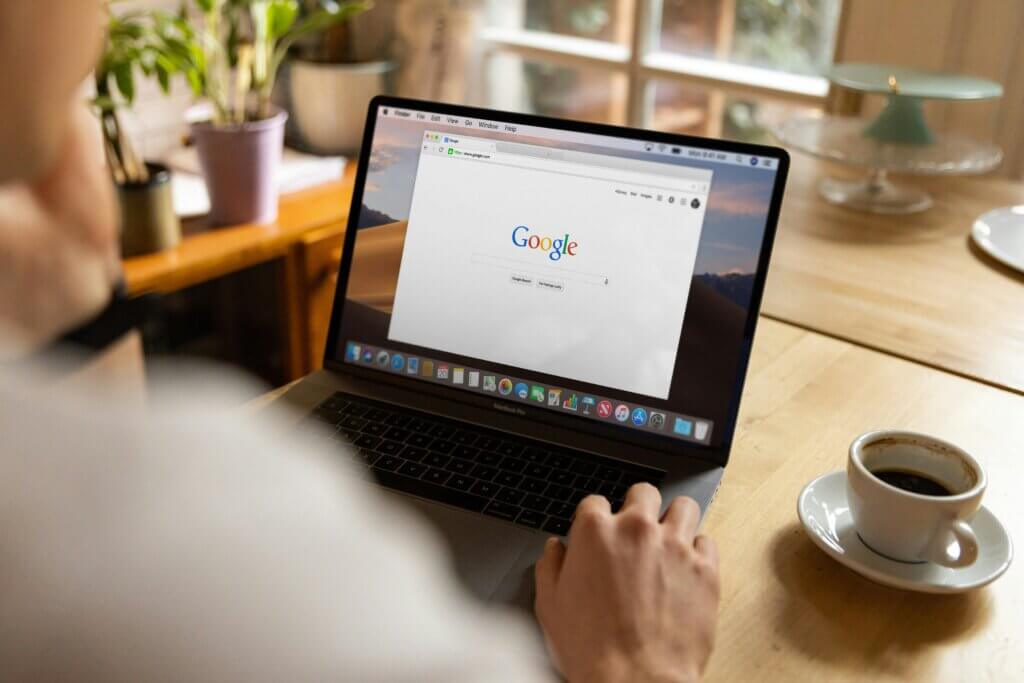 Person sits at light wood table with an espresso cup and saucer and laptop in front of them. The laptop is open to Google and the person's hand hovers near the keyboard as though they're about to search for something.