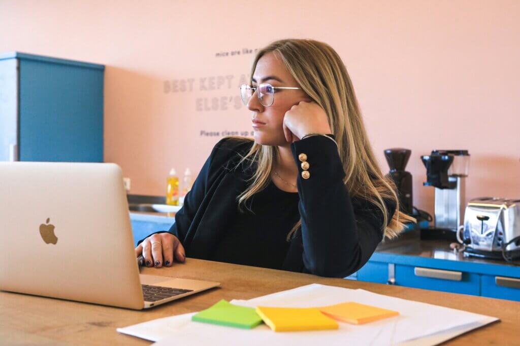 White feminine-presenting person wearing silver glasses, black shirt, and black blazer with gold accents sits at a wooden countertop in a breakroom, with coffee appliances, a sink, and a toaster behind her. The countertop holds paper, sticky notes, and a laptop. She seems to be daydreaming out the window, maybe thinking of a more fulfilling career. 