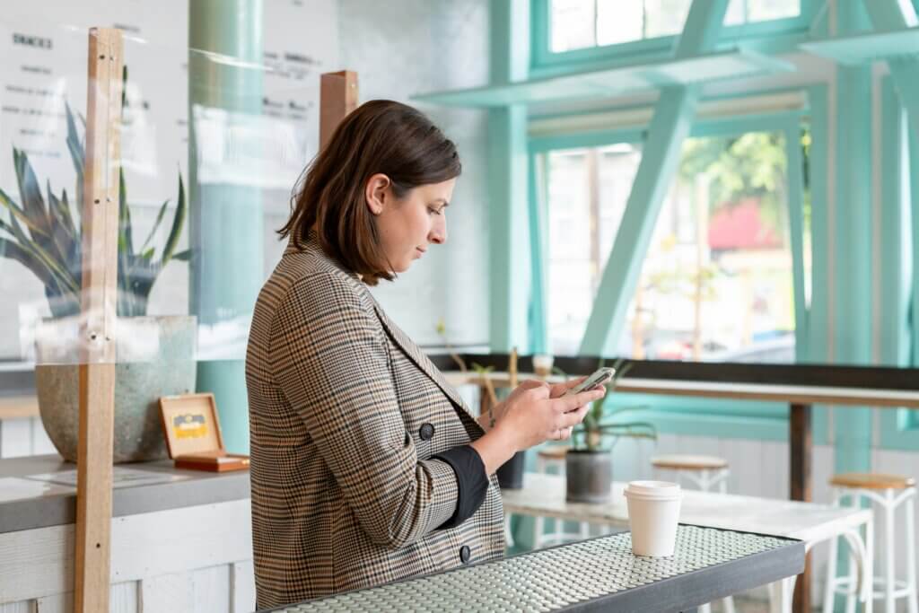 Feminine-presenting person with short brown hair wears a beige plaid blazer and looks down at her phone in her hand. A white to-go coffee cup is on the counter next to her as she stands in a brightly lit turquoise coffee shop.