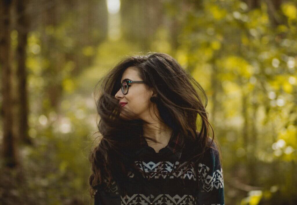 Feminine-presenting person with full dark brown hair quickly turns their head, which makes their hair hang voluminously in the air. They wear black glasses, red lipstick, and a black sweatshirt with white pattern