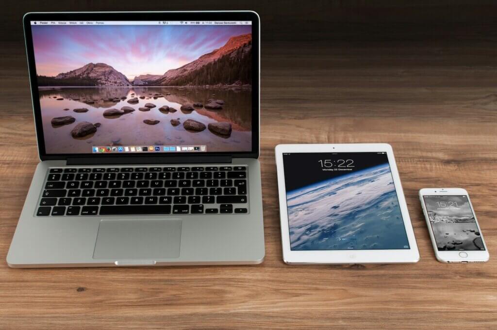 A woodgrain desk with an open Apple MacBook, an iPad tablet, and an iPhone lined up next to each other and all with screens turned on