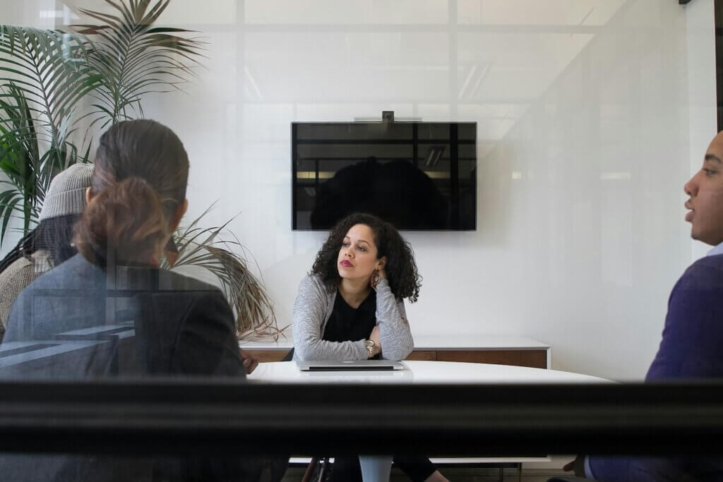 A Black woman with shoulder-length curly hair sits at a white conference table in a meeting with 3 colleagues. She leans on the table and appears to be looking out the window, bored or daydreaming of a dream job while the meeting goes on around her