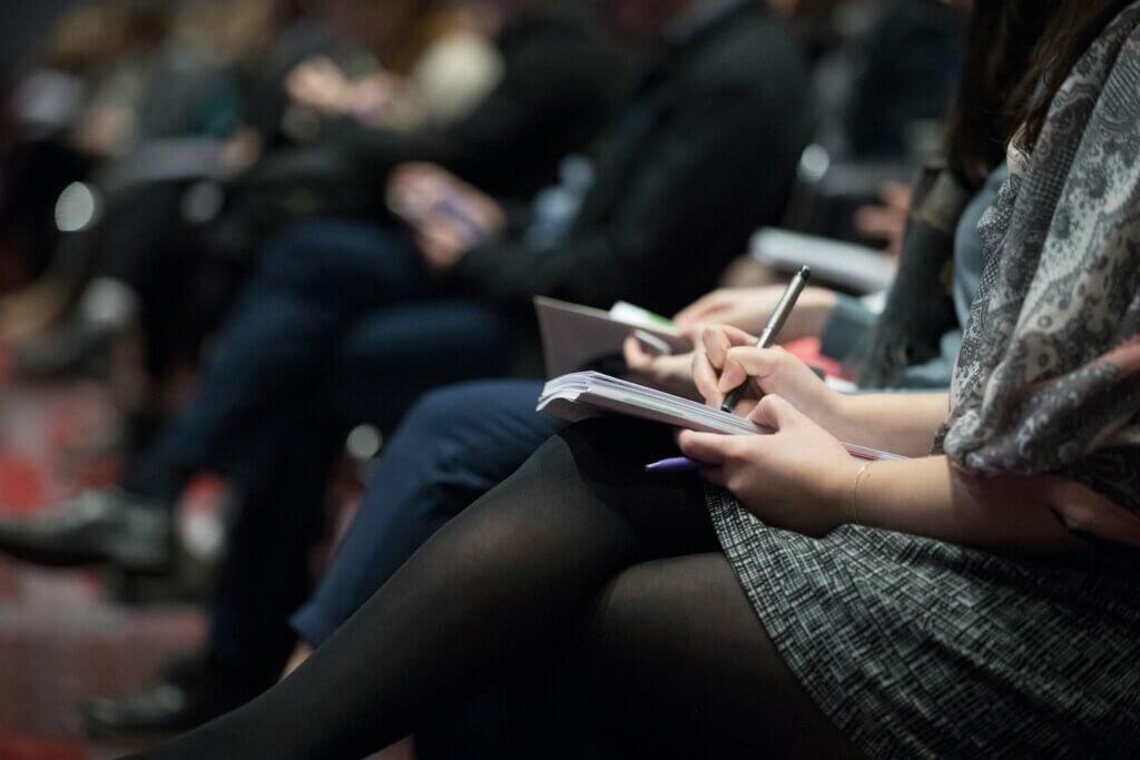 A side view of the lap of a business woman in gray skirt and black pantyhose or tights as she takes notes in a seminar or educational class or session. Out of focus in the background is the rest of the row of others in the class.