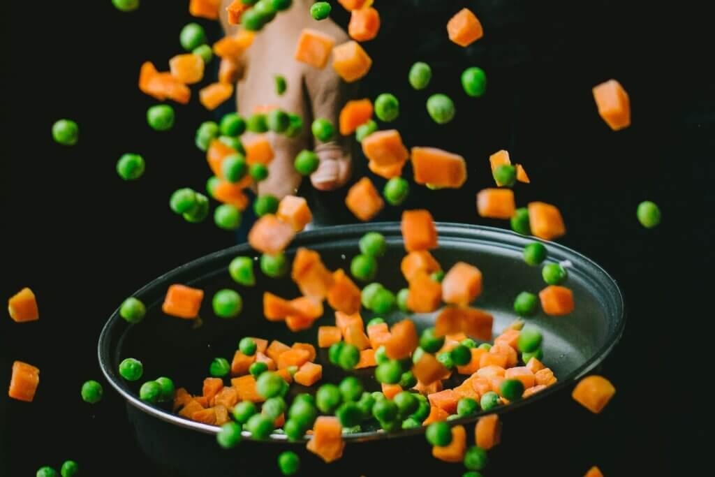 A person of color holds a frying pan and flips a bunch of chopped carrots and peas, which scatter midair in the picture