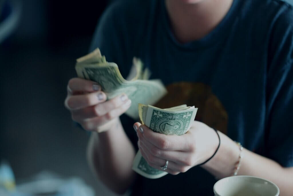 A white person wearing a dark blue t-shirt counts a stack of $1 bills to save money