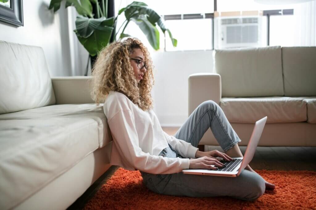 Feminine-presenting person of color with shoulder-length curly blonde hair sits on a red rug in an apartment setting, leaned up against a white leather couch. She wears a white turtleneck and cropped light blue jeans and is working on a laptop balanced on one knee.