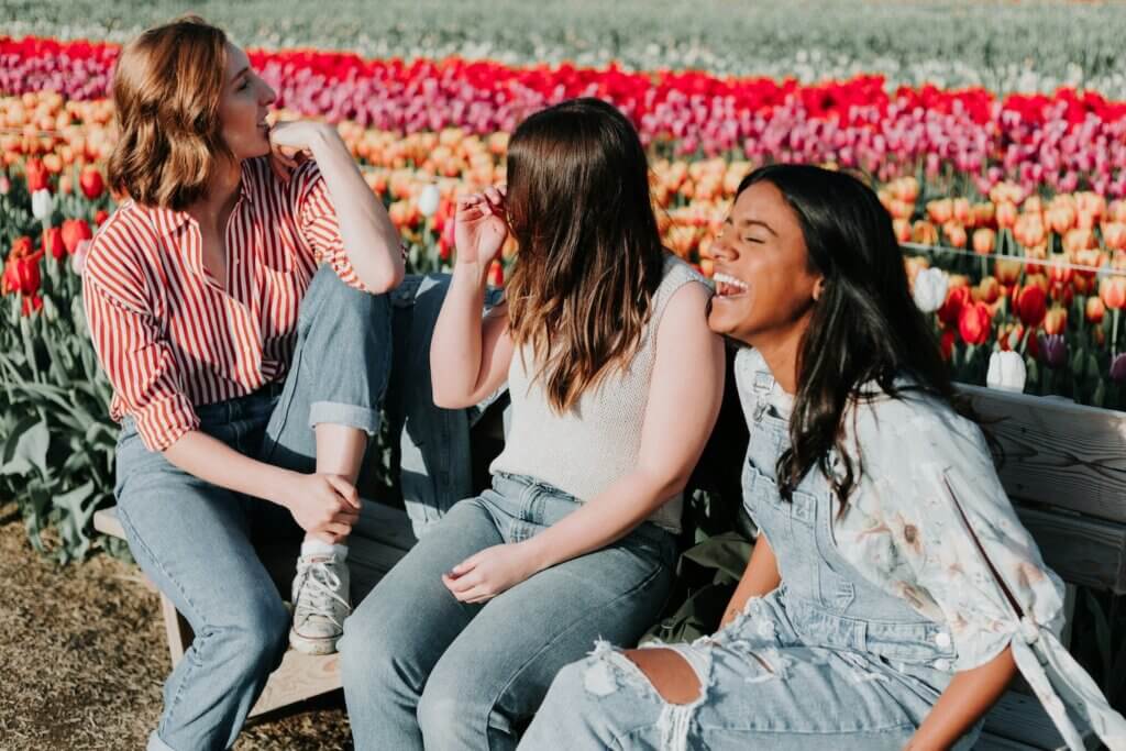 Three feminine-presenting people, two white and one brown-skinned, sit on a bench in a field of tulips. It's a sunny day, and they're laughing together, representing a supportive network of friends