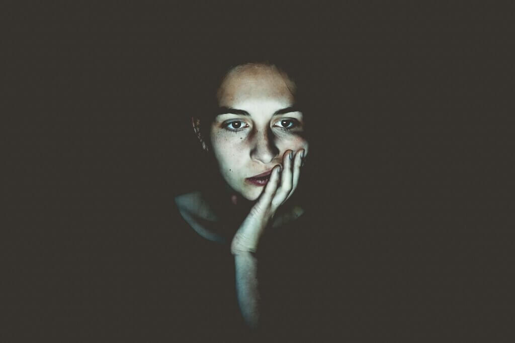 In a dark room with totally black background, the face of a white feminine-presenting person sits illuminated by the glow of a computer screen. She appears to be concentrating on something, with her hand on her face, but she also looks bored and may be thinking, "I don't want a career!"