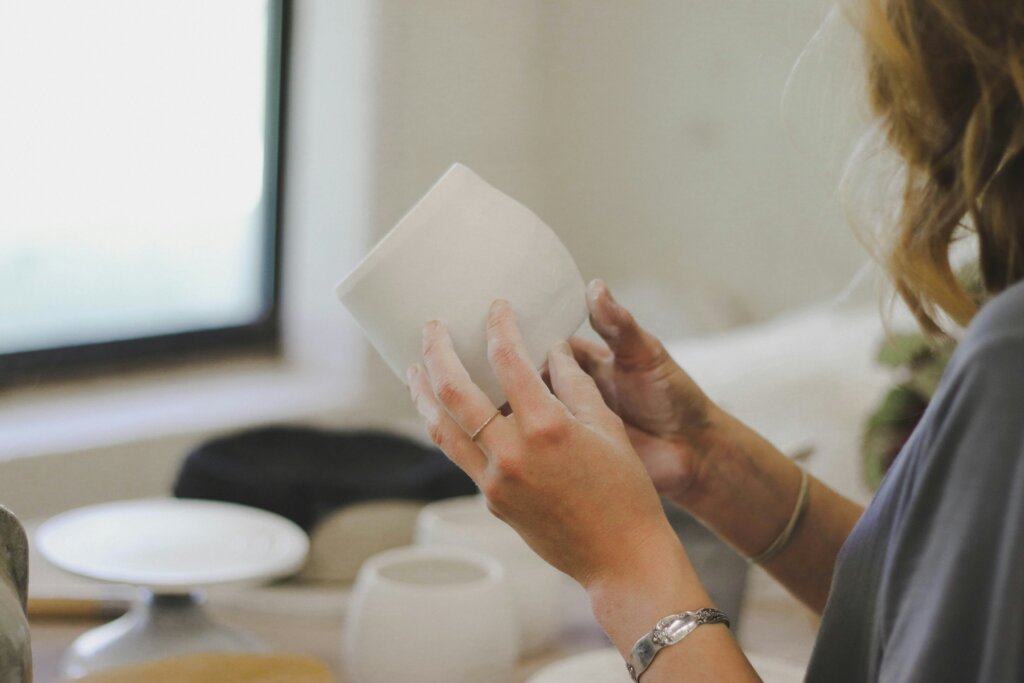 A white person with blonde wavy hair holds a white piece of pottery in a pottery studio. Their face isn't visible. They have a ring on their left middle finger and a silver bracelet on each wrist. The image represents a person with no interest in their career pursuing hobbies