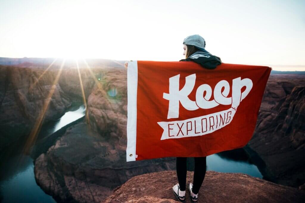 A woman in gray baseball cap, black pants, and black sandals stands at the top of a red rock cliff with a lake below. She extends her arms to hold a large red flag wrapped behind her; the flag reads "Keep exploring" and represents a journey mindset for your career discovery process