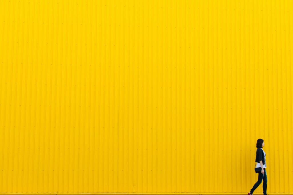 A white woman with dark hair wears black and white clothes and walks in front of a huge industrial wall painted bright yellow