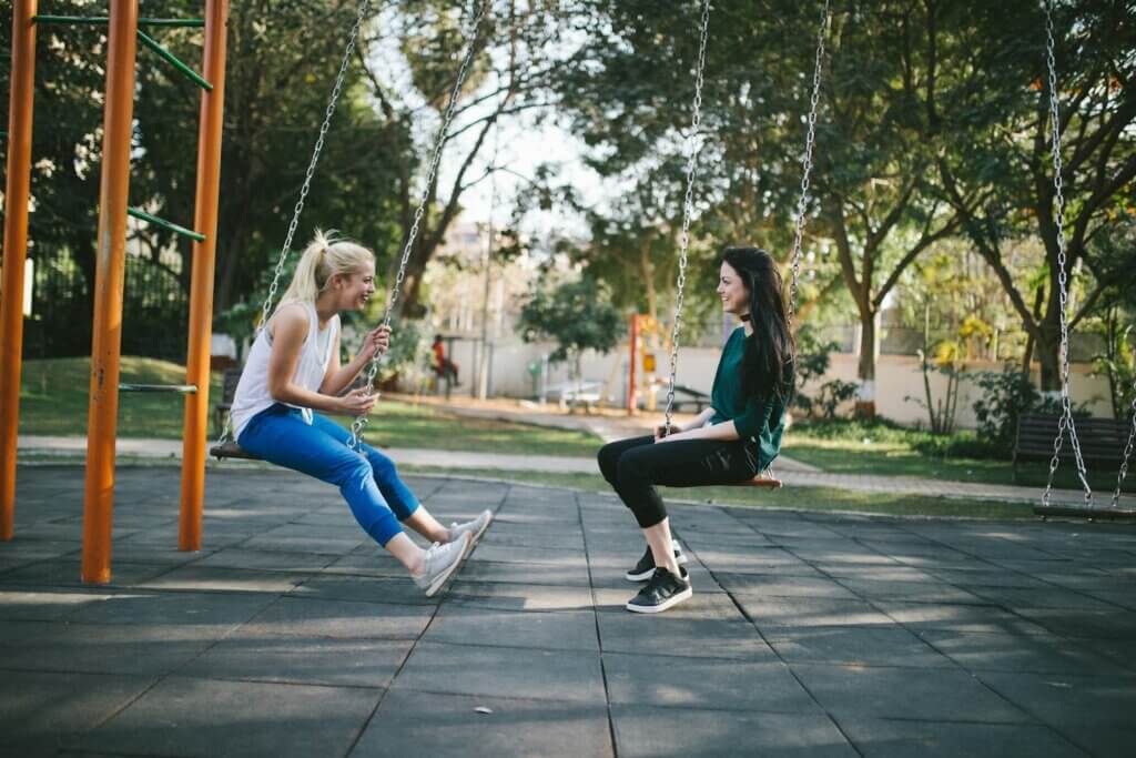 Two white women laugh and swing on swings in a park. They might be celebrating a good workout or just hanging out. One has bleached blond hair and wears a white shit with blue leggings; the other has dark brown hair and wears a green shirt with black leggings