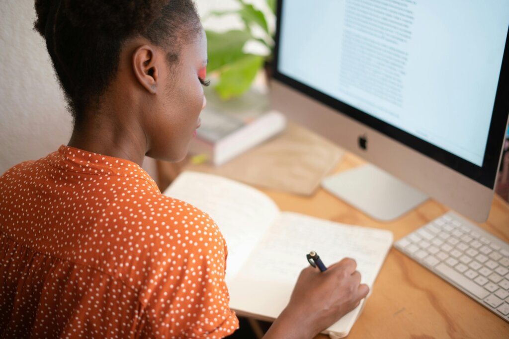 Black feminine person in orange shirt with white dots writing in notebook. They sit at a wooden desk with an Apple desktop computer and houseplant out of focus.