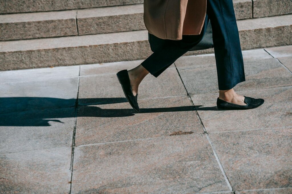 The legs and feet of a feminine person in beige coat and black flats walking on a tiled sidewalk