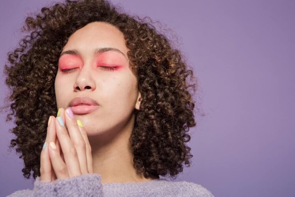 A feminine person of color with curly hair and bright neon pink eyeshadow has eyes closed and hands up to her face, as though thinking through something in depth (like career confusion!). She wears a light purple sweater.
