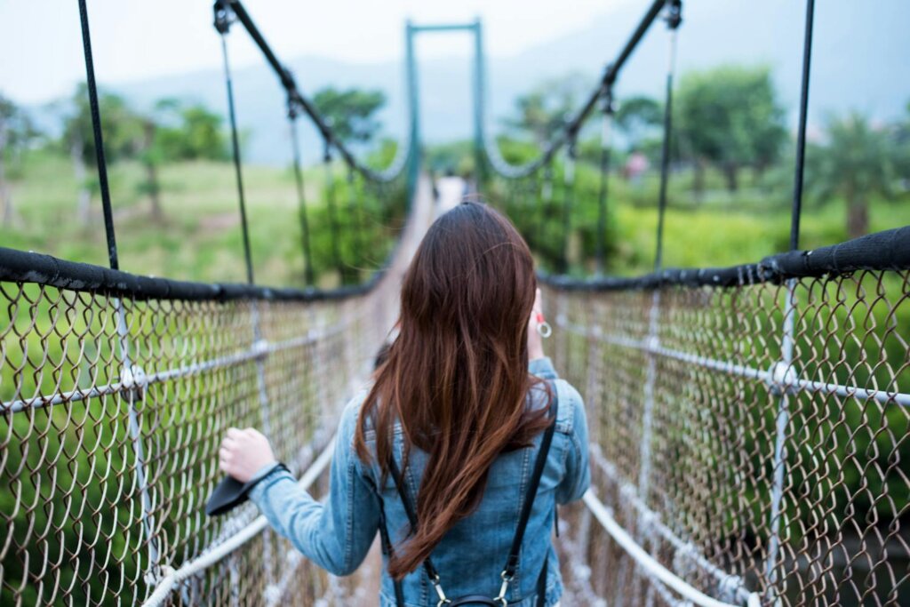 feminine person with long dark hair wearing jean jacket on rope bridge surrounded by lush green grass, trees, and mountains in the distance