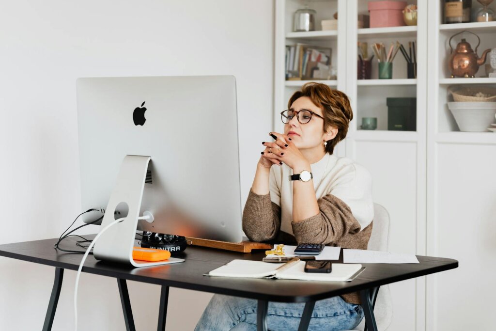 Feminine white person with short light brown hair and glasses sits at a desk with an Apple desktop computer. Elbows rest on the desk as she's thinking about something, looking off to the side