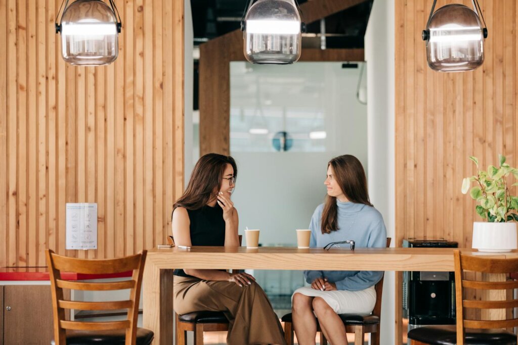 two feminine-presenting people chatting in coffee shop; on the left is an Asian person with brown hair, black sleeveless top, wide light brown pants, and glasses. On the right is a white person with long brown hair, blue turtleneck, and white skirt.
