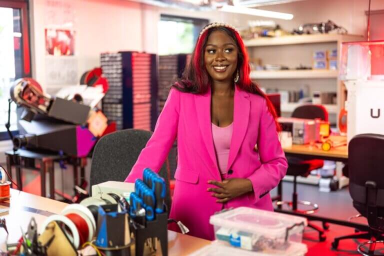 Smiling female engineer in pink suit standing in a busy workshop surrounded by tools and electronics.