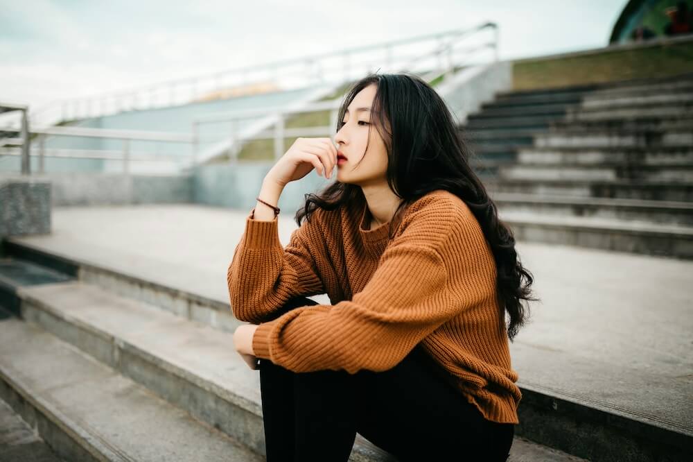 A young Asian feminine-presenting person with fair skin and long black hair sits on empty bleachers, with one arm propped up on her knee and fingers grazing her lips. She looks lost in thought. She wears a brown sweater and black pants.