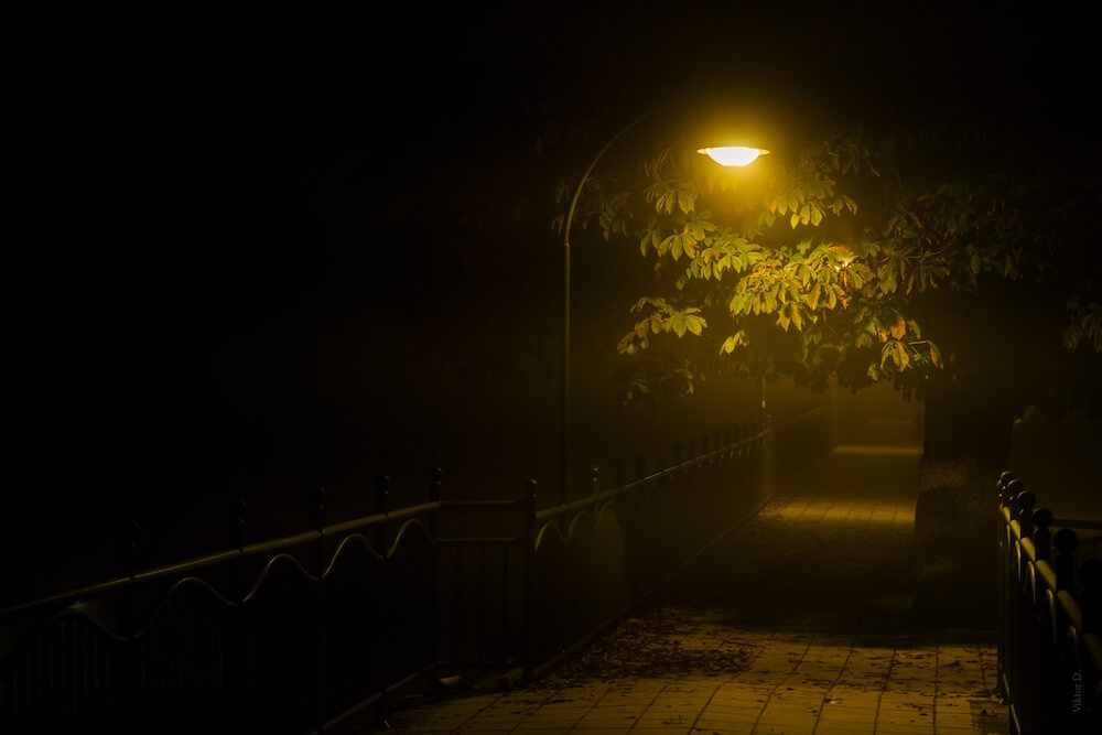 A dark path or bridge at night, with iron railings on either side and a single street lamp partially illuminating the path