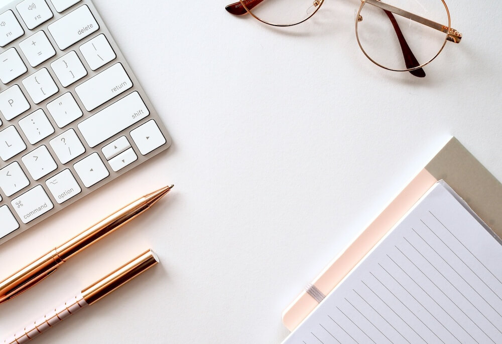 A white desktop with an Apple keyboard, rose-gold pens, round wire glasses, and lined paper notebooks
