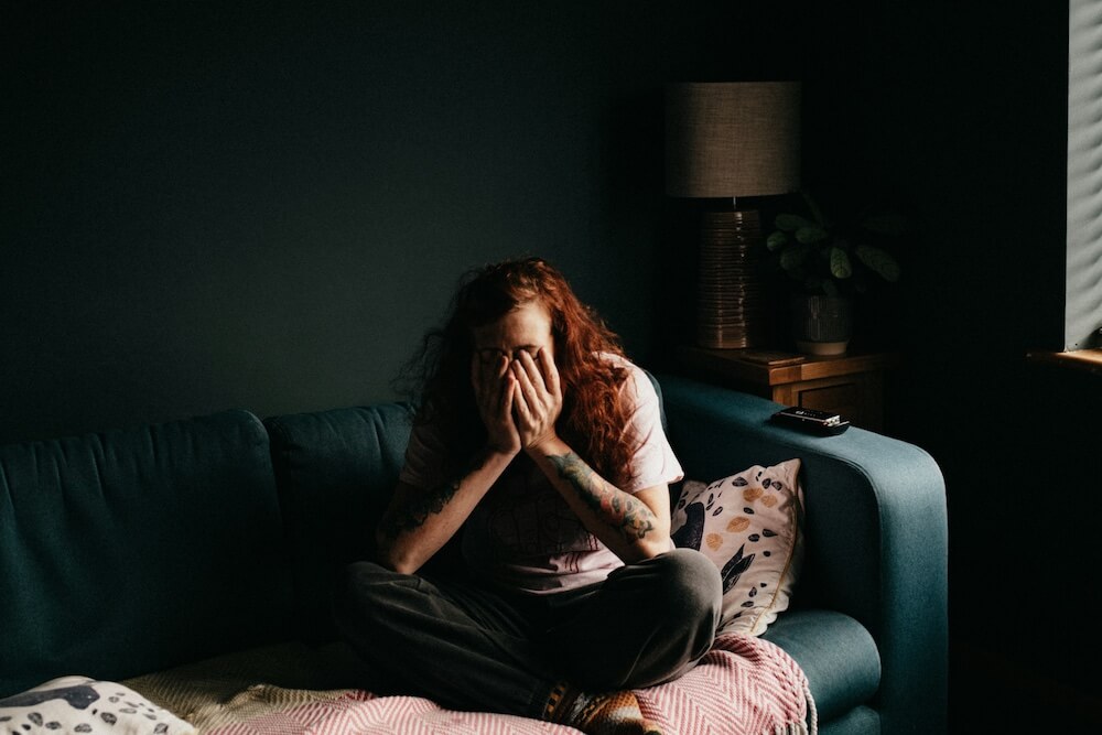 A young feminine-presenting person with long wavy natural red hair sits cross-legged on a teal couch in a dark green room, her hands held up to her face. Her posture looks stressed, and we can't see her face.
