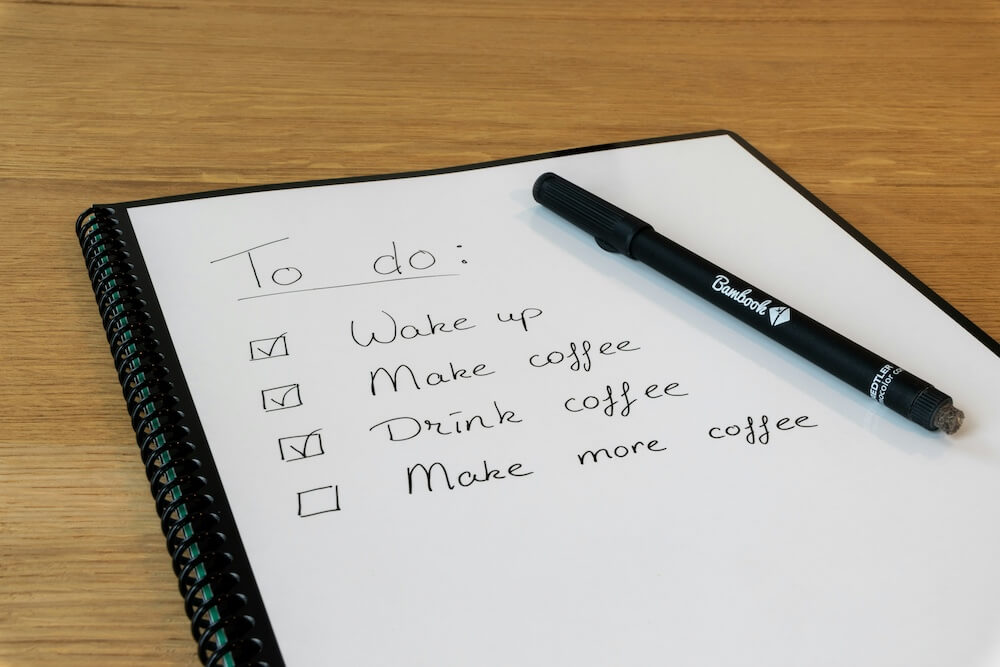 a notebook with a page of checklist items checked off, including wake up, make coffee, and drink coffee. The next unchecked item on the list is "drink more coffee"